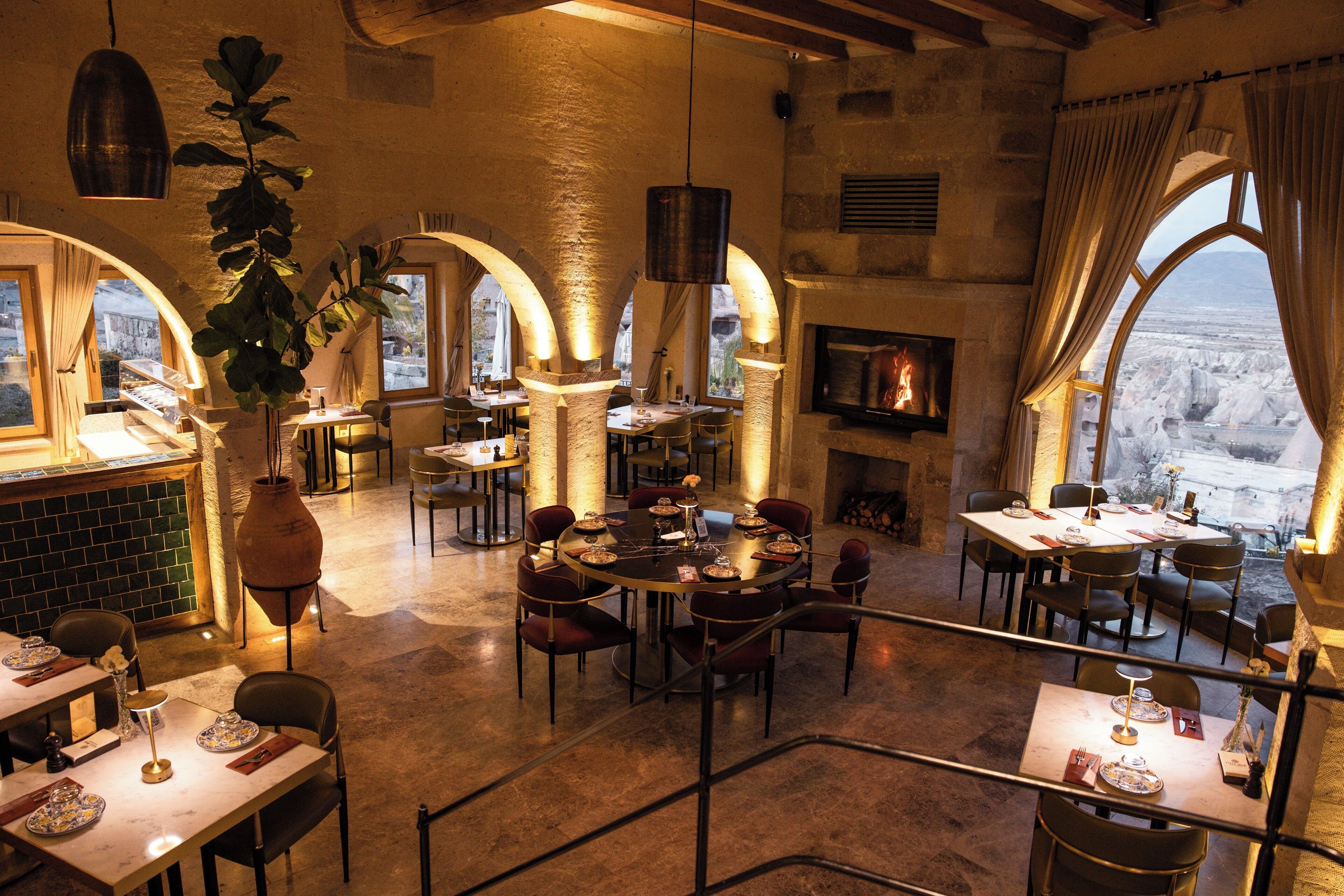 A restaurant with multiple tables and chairs, a fireplace, and large windows with a view of a snowy landscape.