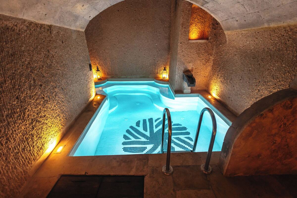 A stone-walled hot tub with a leaf mosaic design, surrounded by stone arches and illuminated by wall-mounted lights.