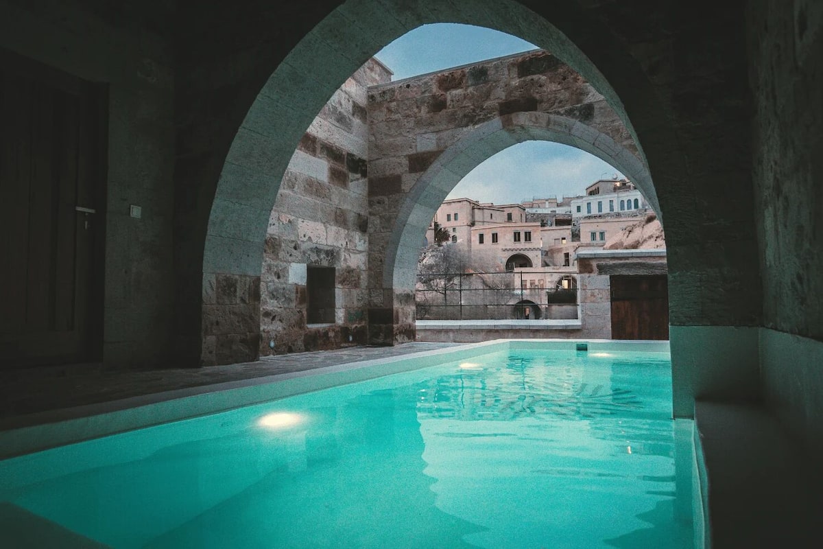 An indoor swimming pool with arched stone walls and a view of historic buildings in the background.