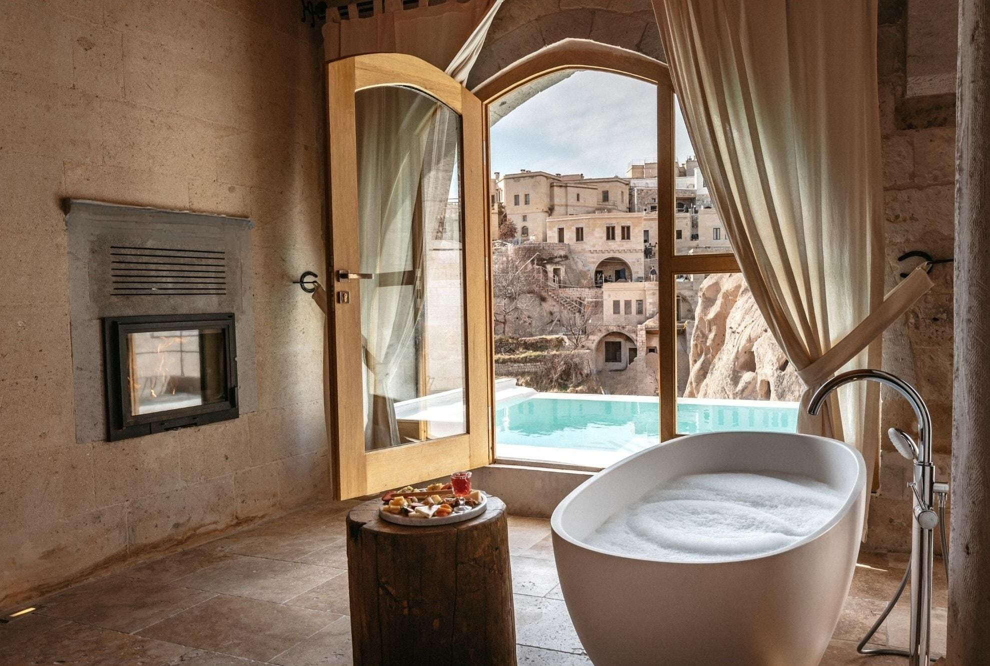 Un baño con una bañera independiente, un taburete de madera con aperitivos, y vistas a una piscina y edificios a través de una ventana.