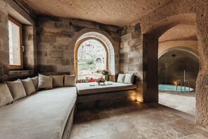 A stone-walled room with a built-in bench, a small table with a vase and fruits, and a view of a pool through an arched window. - Millstone Cave Suites (Nevsehir)
