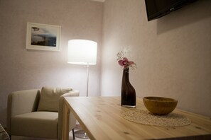 A room with a wooden table, a vase with flowers, a wooden bowl, a lamp, and a television on the wall.