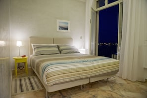A bedroom with a bed, a nightstand, a lamp, a picture on the wall, and a view of the night sky through a window. - Garibaldi R&B (Messina)