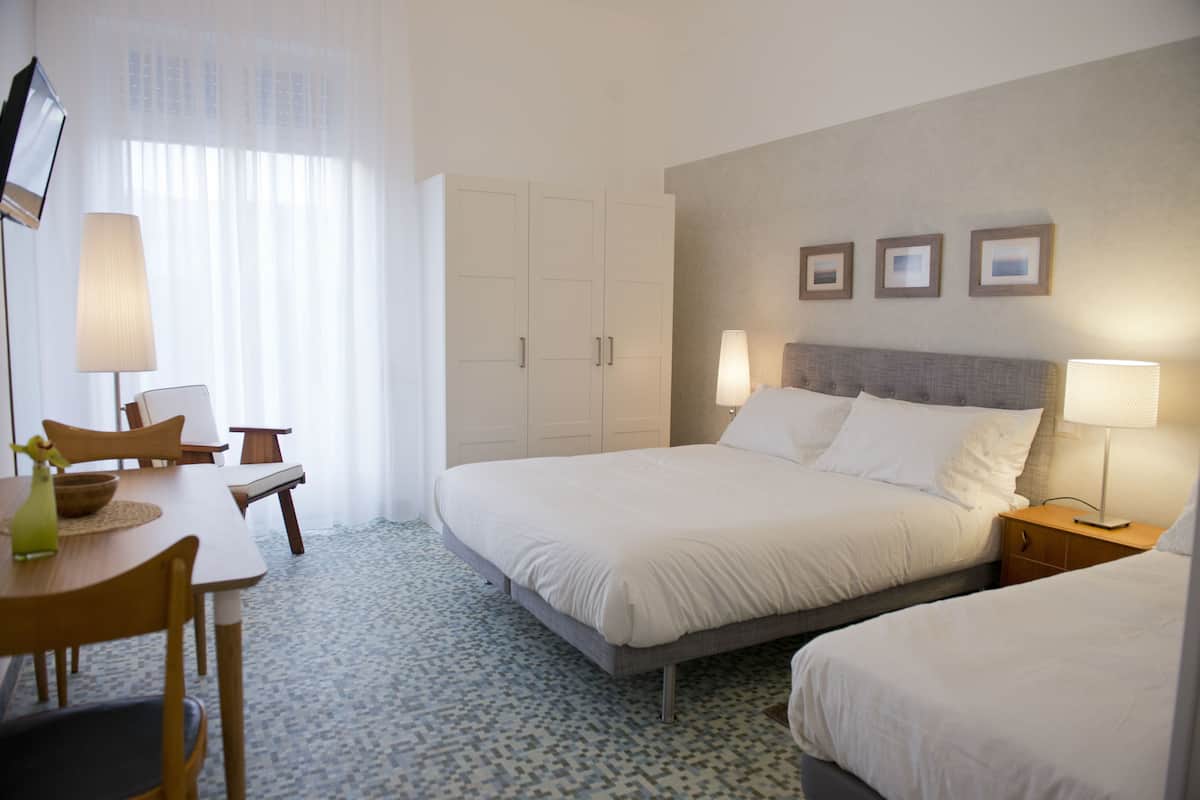 A hotel room with a large bed, a desk with a chair, a small table with a vase, and a lamp. There are three framed pictures on the wall above the bed.