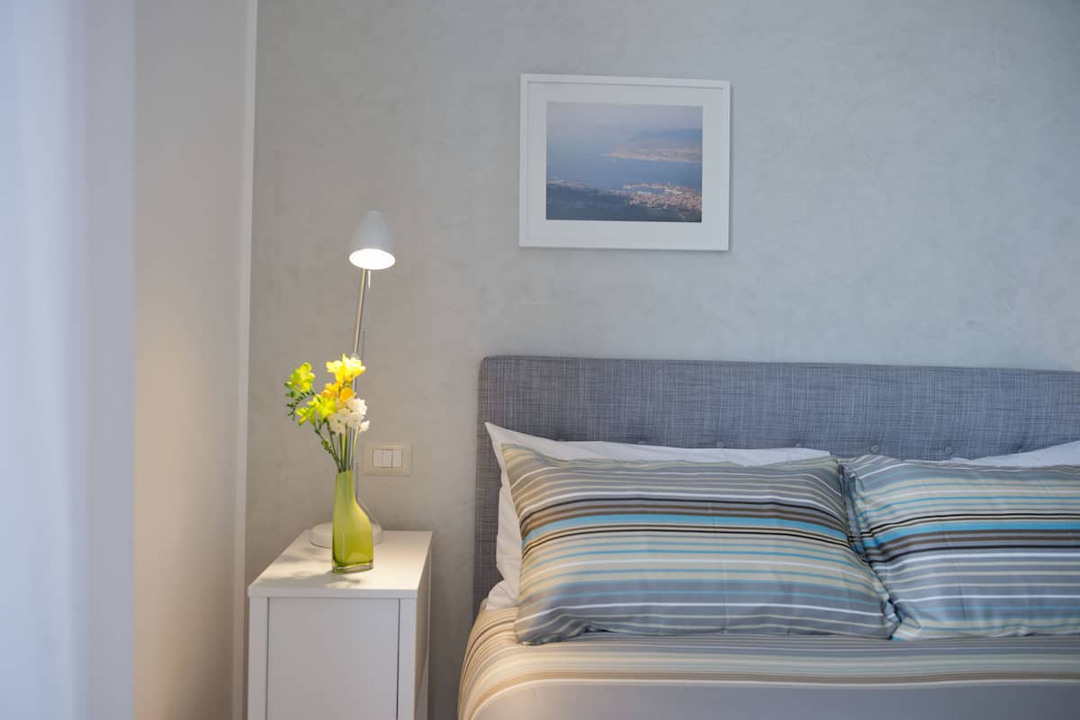 A neatly made bed with striped pillows, a bedside lamp, and a vase with flowers on a bedside table.