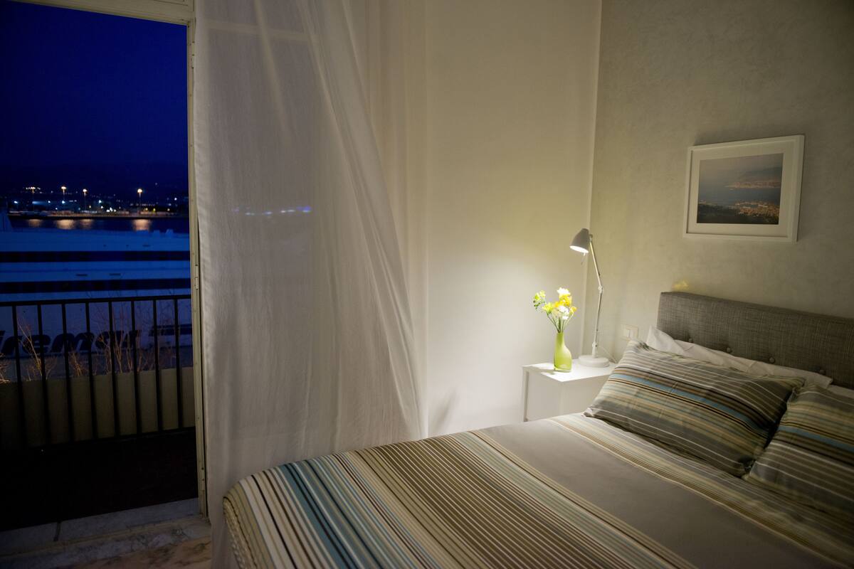A bedroom with a bed, a nightstand, a lamp, a vase with flowers, a framed picture on the wall, and a view of the water outside.