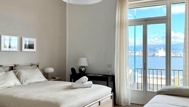 Ett hotellrum med en stor säng, ett skrivbord, en stol, en lampa och utsikt över en sjö eller ett annat vattendrag genom stora fönster.
