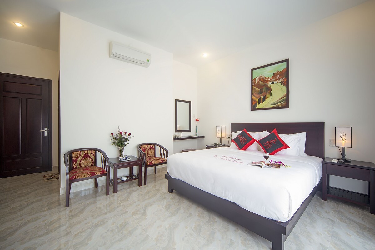 Une chambre d’hôtel avec un grand lit, deux chaises, une petite table, une table de chevet, un climatiseur fixé au mur, un tableau encadré et une porte.