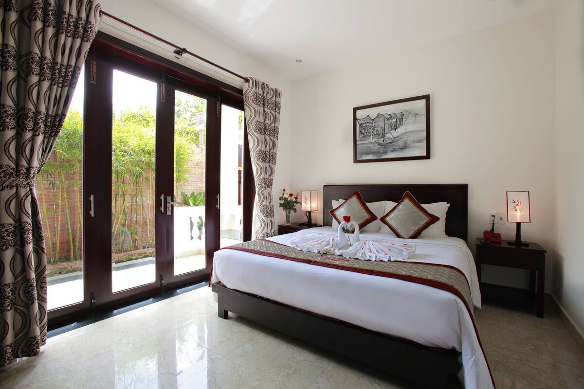 Une chambre d’hôtel avec un lit, une table de chevet avec une lampe, un tableau au mur et une vue sur la verdure à l’extérieur.