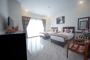 Quarto de hotel com duas camas, televisão, mesa de jantar com cadeiras e uma janela grande com cortinas.