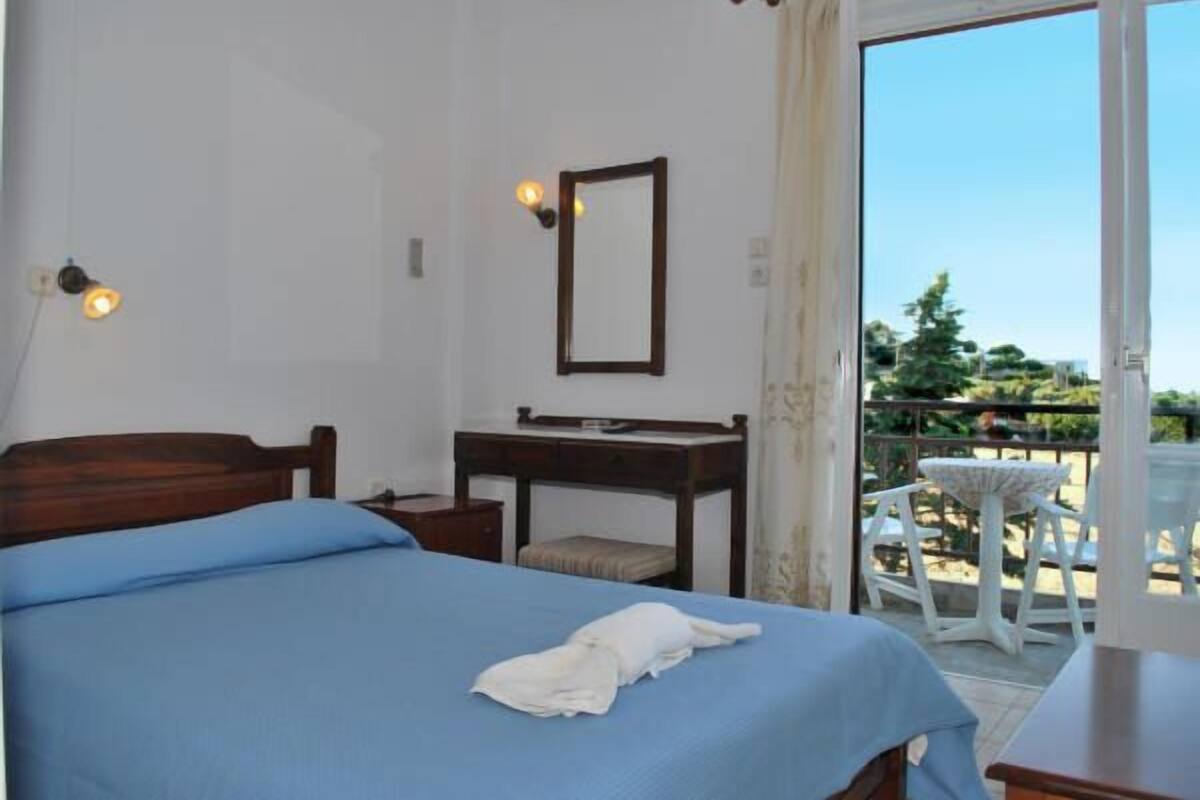 A bedroom with a bed, a desk, and a chair. There is a balcony with outdoor furniture.