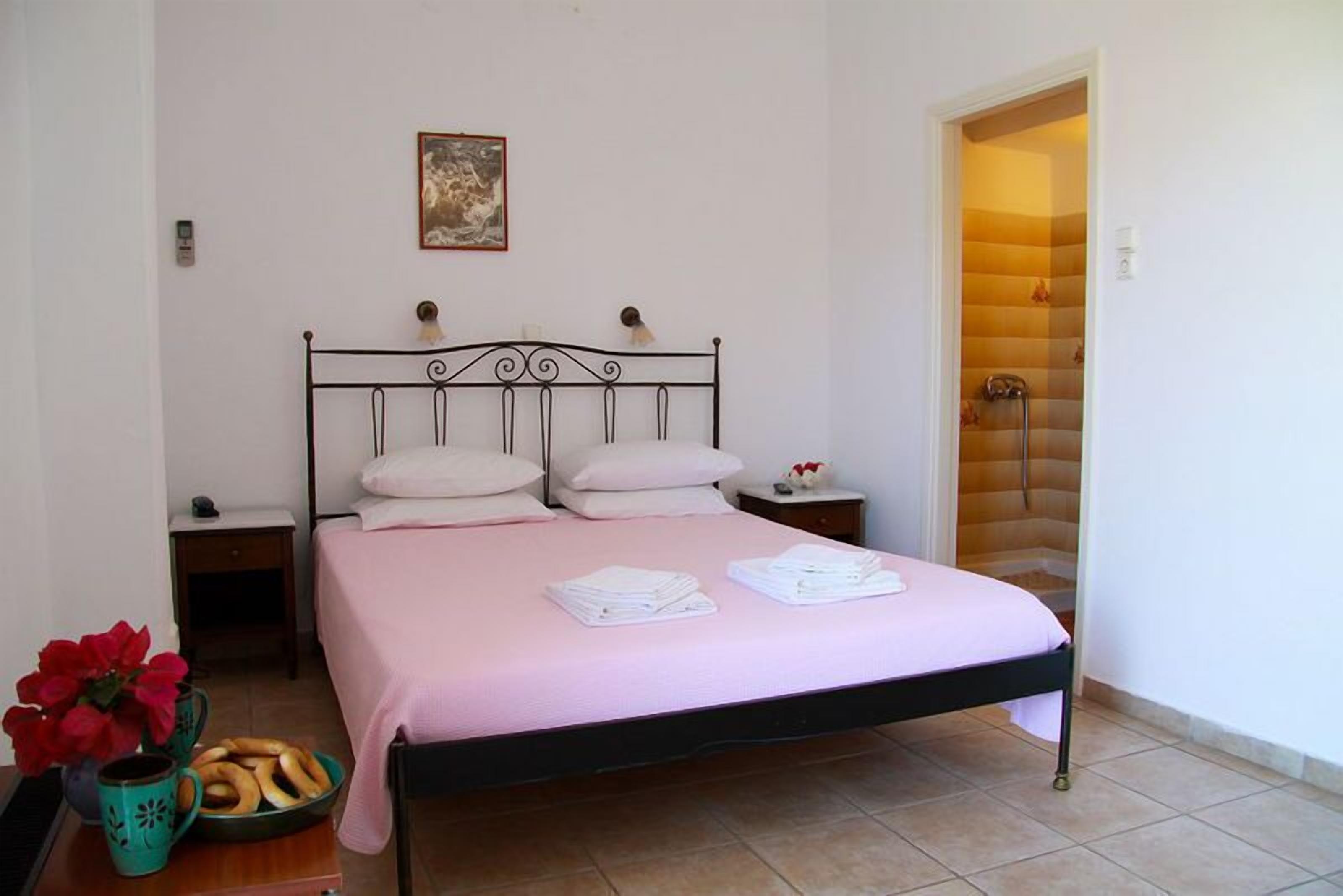 Una cama con ropa de cama rosa, dos almohadas y toallas dobladas sobre una estructura metálica, en una habitación con paredes blancas y piso de baldosas.