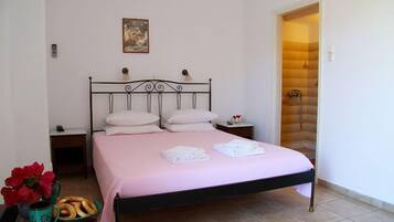 Una cama con ropa de cama rosa, dos almohadas y toallas dobladas sobre una estructura metálica, en una habitación con paredes blancas y piso de baldosas.