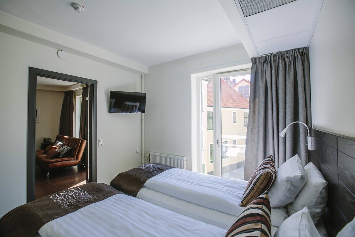 Quarto de hotel com duas camas, televisão, janela com cortinas e vista para edifícios.