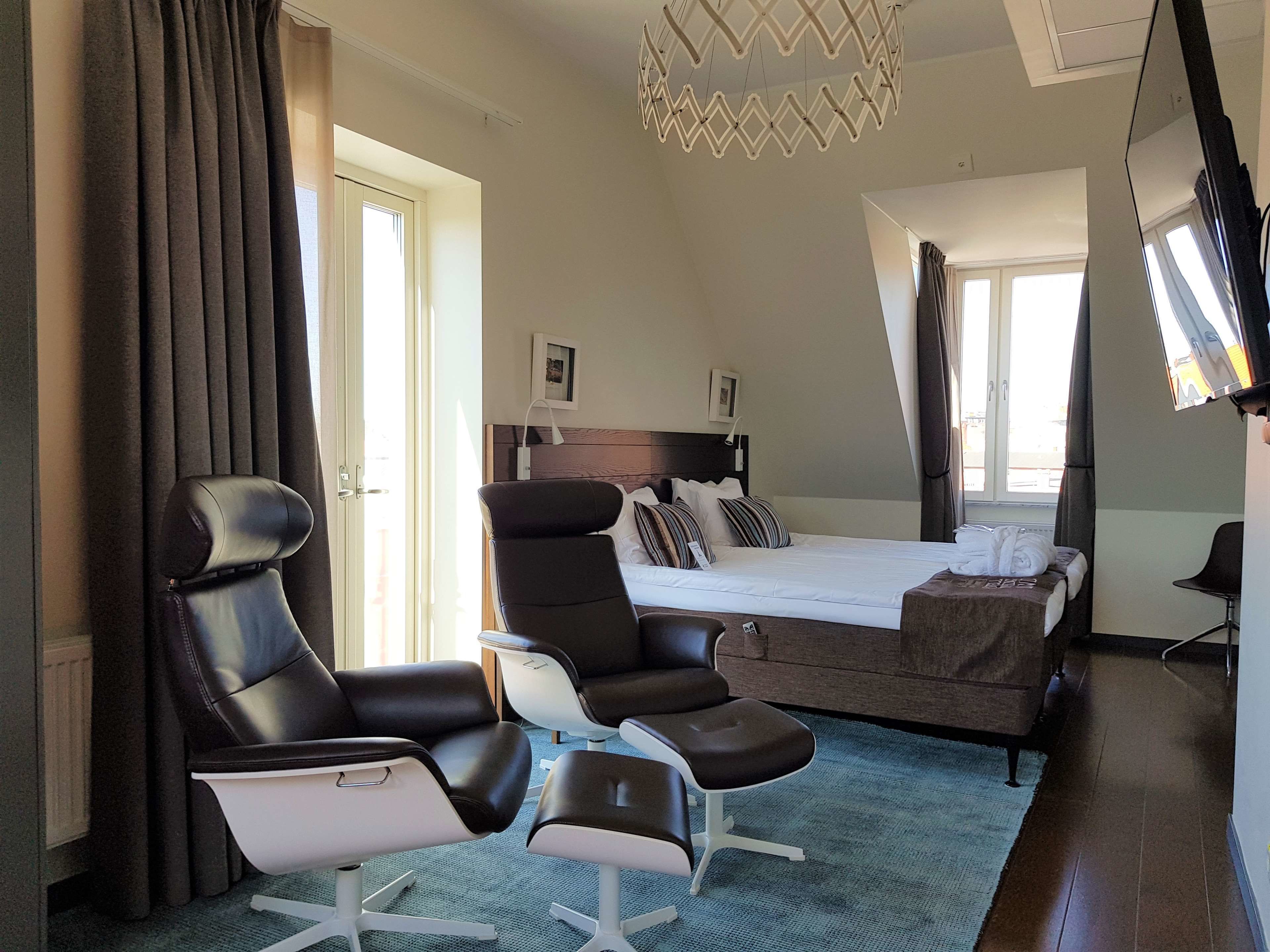 Una habitación de hotel con una cama, dos sillones reclinables, un televisor y un ventanal grande.
