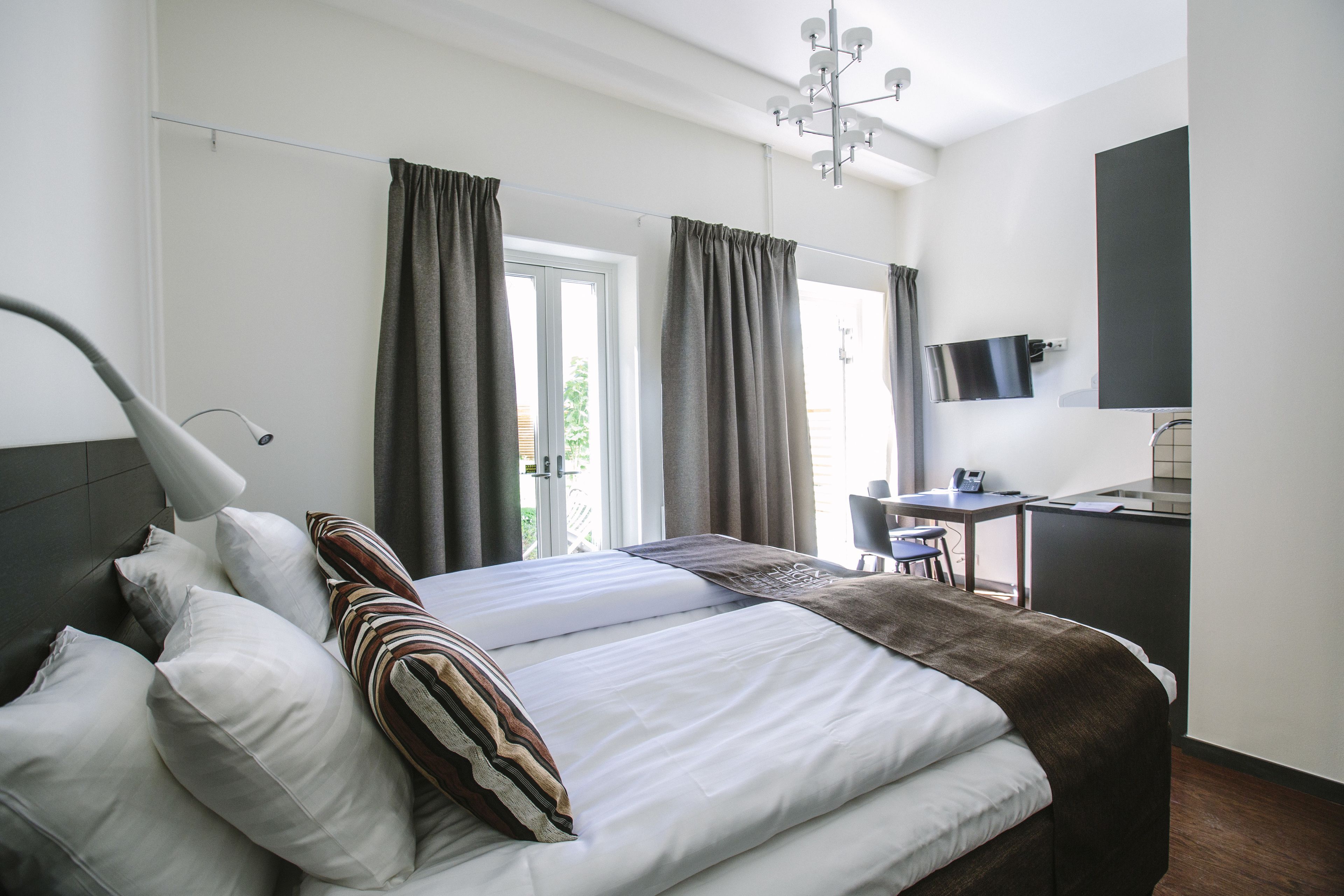 Una habitación de hotel moderna con una cama bien hecha, un escritorio con silla, un televisor y un ventanal con cortinas.