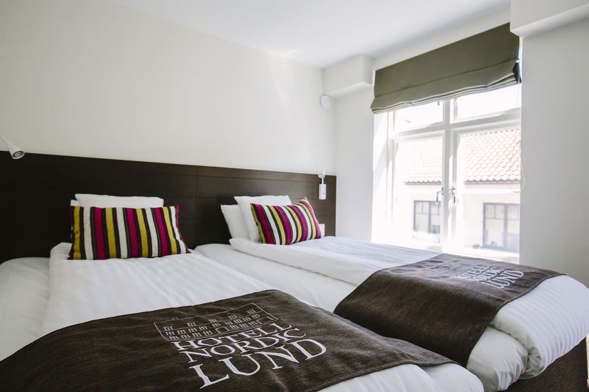 Quarto de hotel com duas camas, cabeceira escura e janela com vista para edifícios.