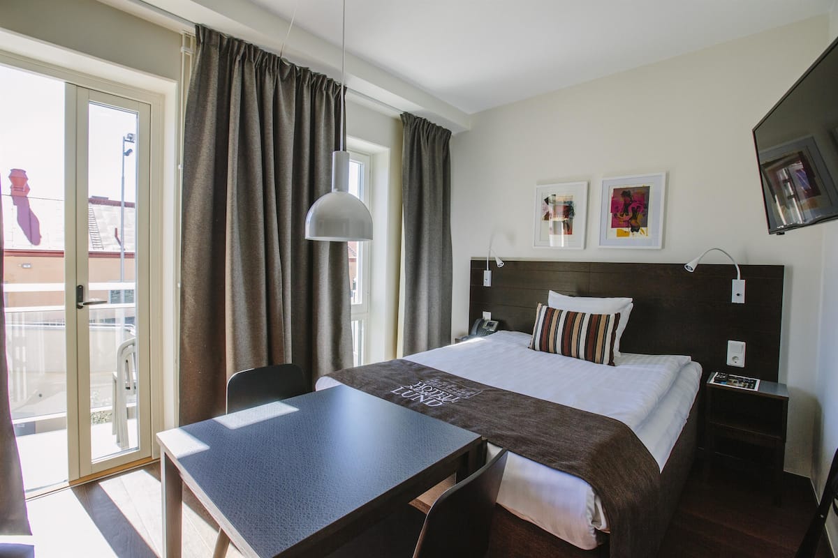 Quarto de hotel com cama, mesa, cadeira, escrivaninha, mesa de cabeceira e janela com cortinas.