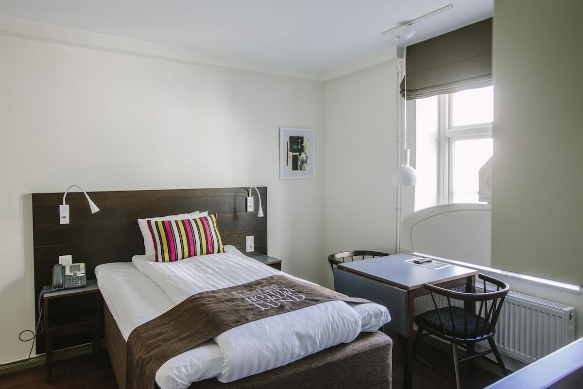 Quarto de hotel com cama, mesa de cabeceira, cadeira, escrivaninha e janela.