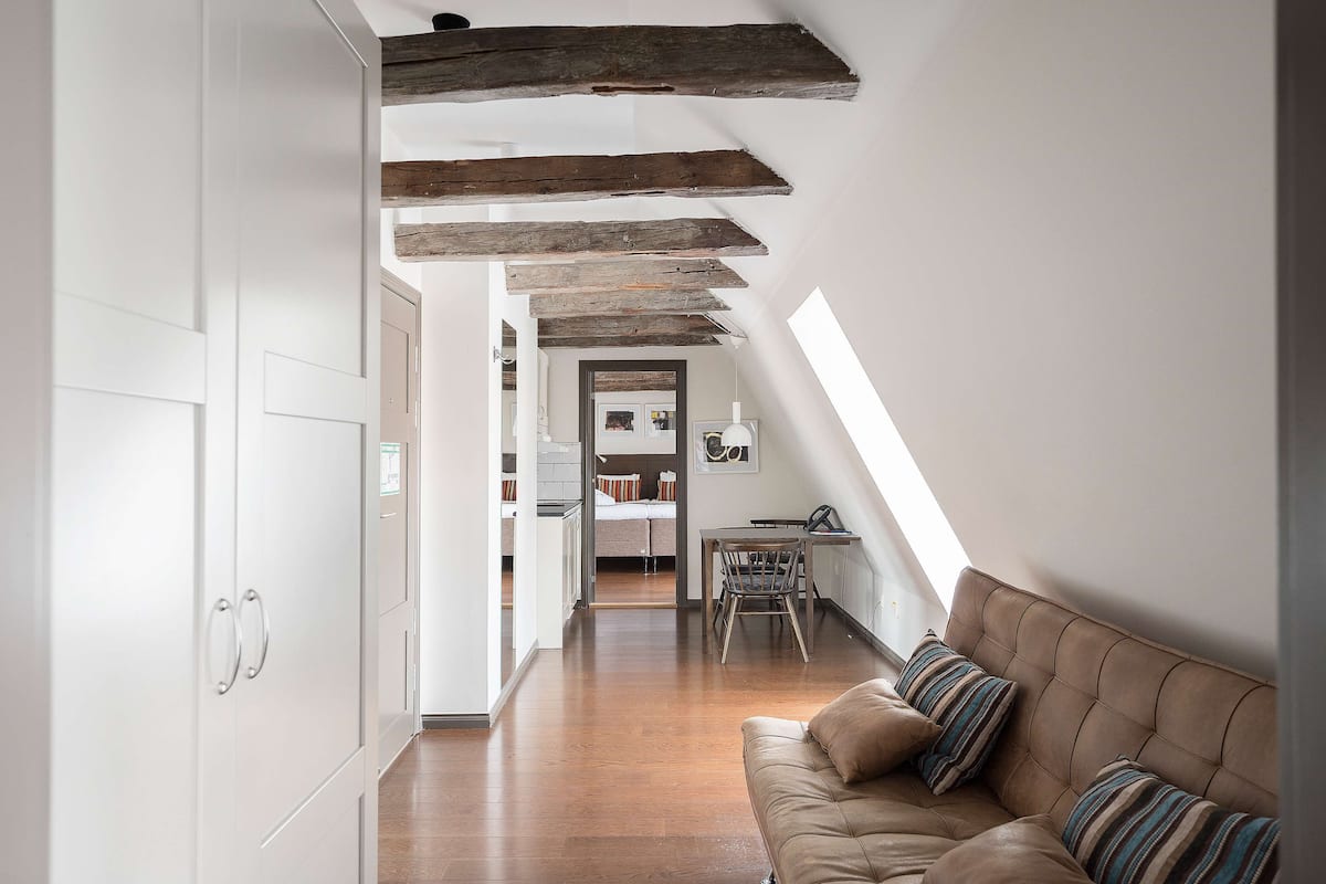 Um loft moderno com vigas de madeira aparentes, um sofá de couro marrom aconchegante e uma área de refeições com uma mesa pequena e cadeiras.
