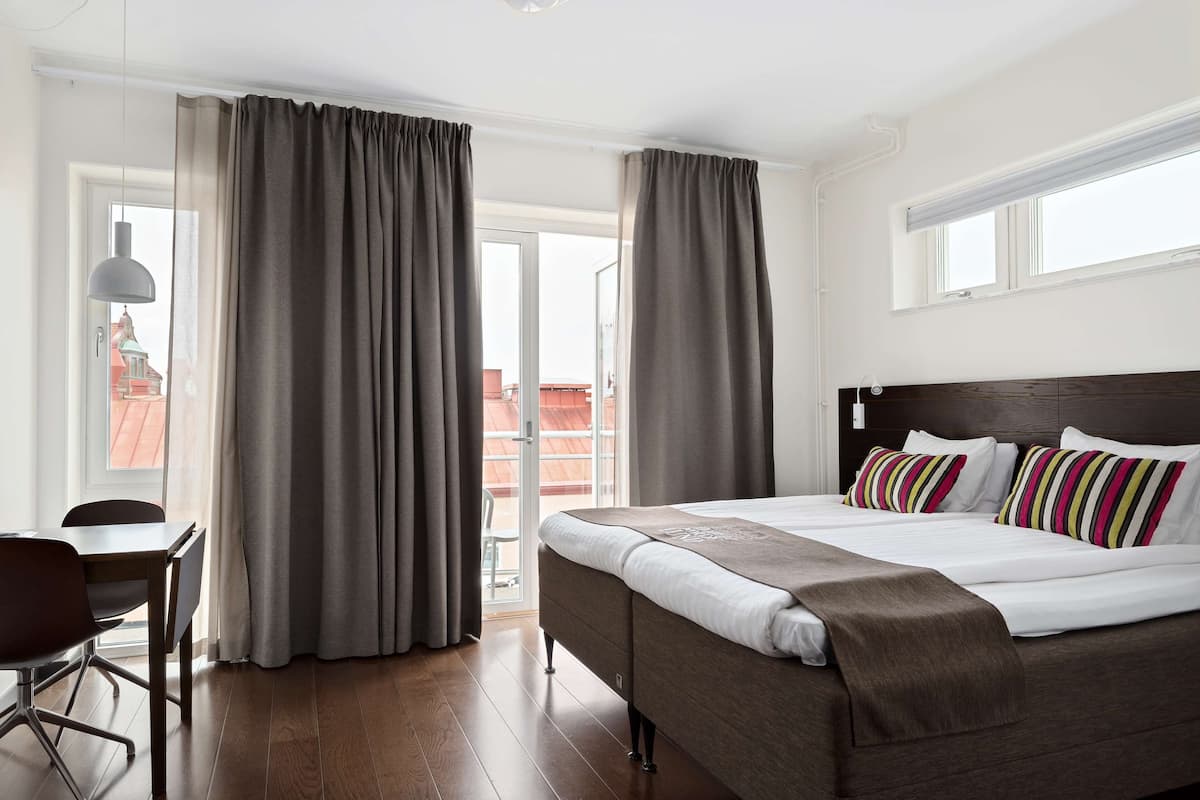 Quarto de hotel com uma cama grande, uma escrivaninha e vista para um edifício com telhado vermelho pela janela.