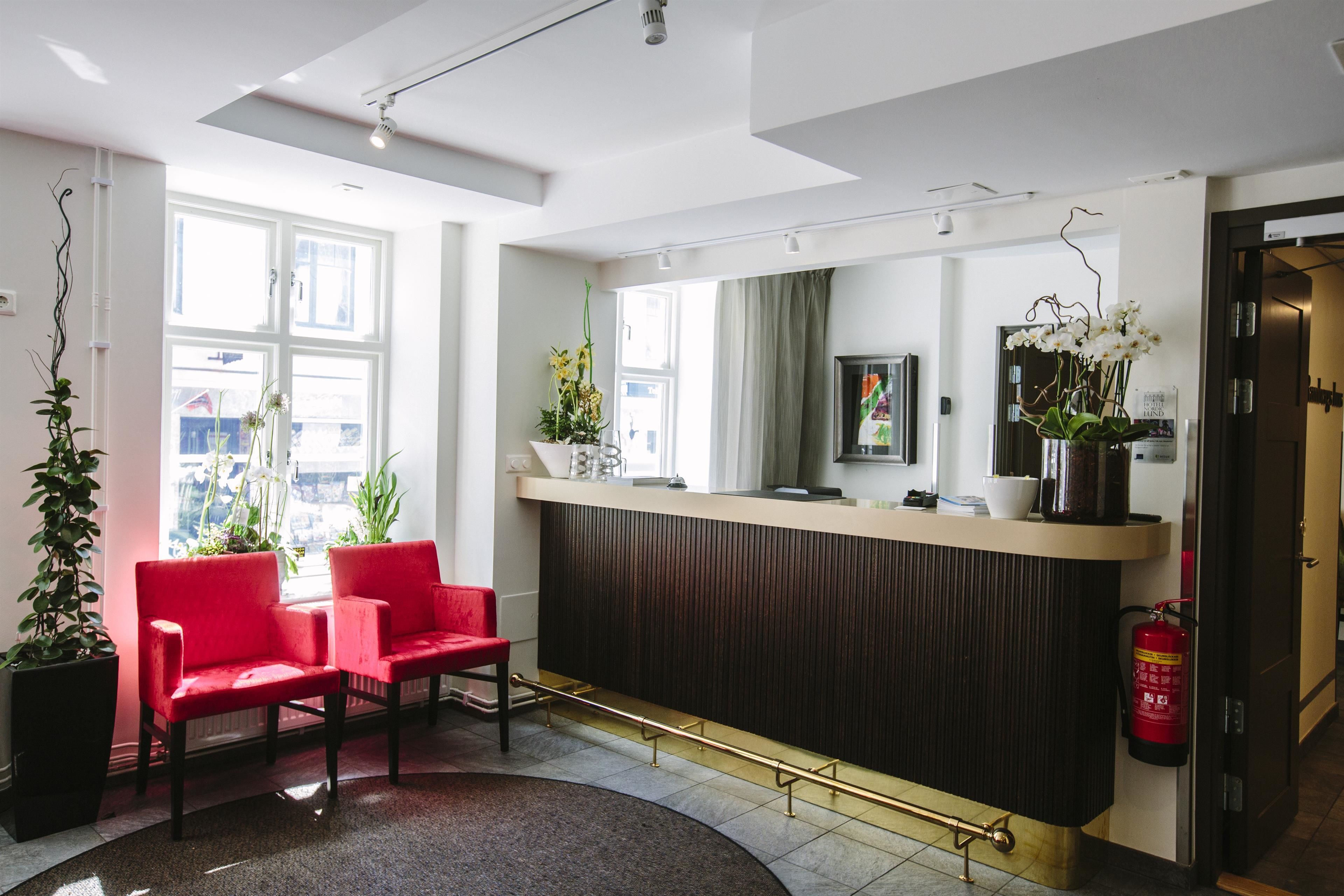 Ett välkomnande område med modern design, med en receptionsdisk, två röda stolar, krukväxter och en brandsläckare.