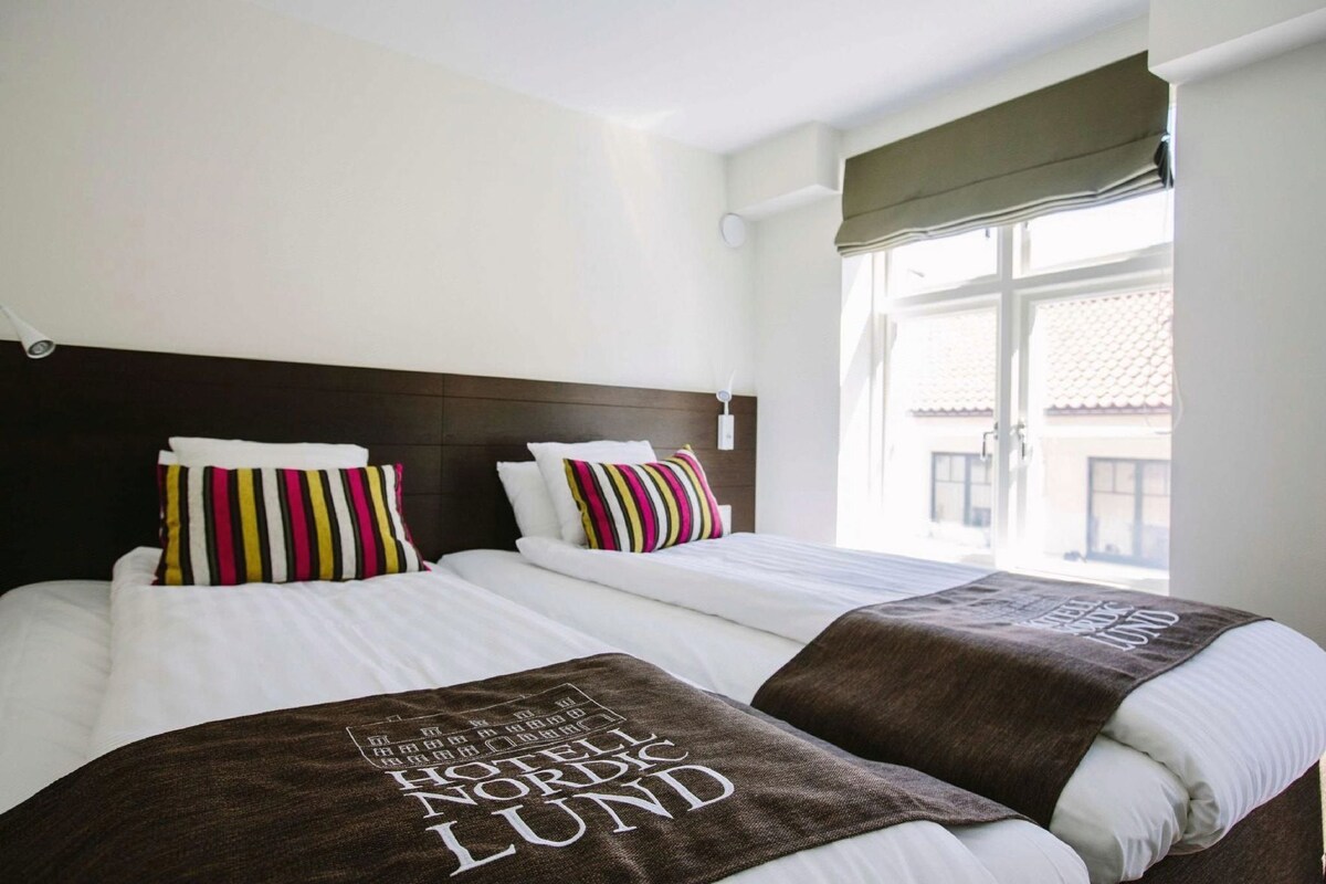 A hotel room with two beds, striped pillows, a window with a view, and a brown blanket with text.