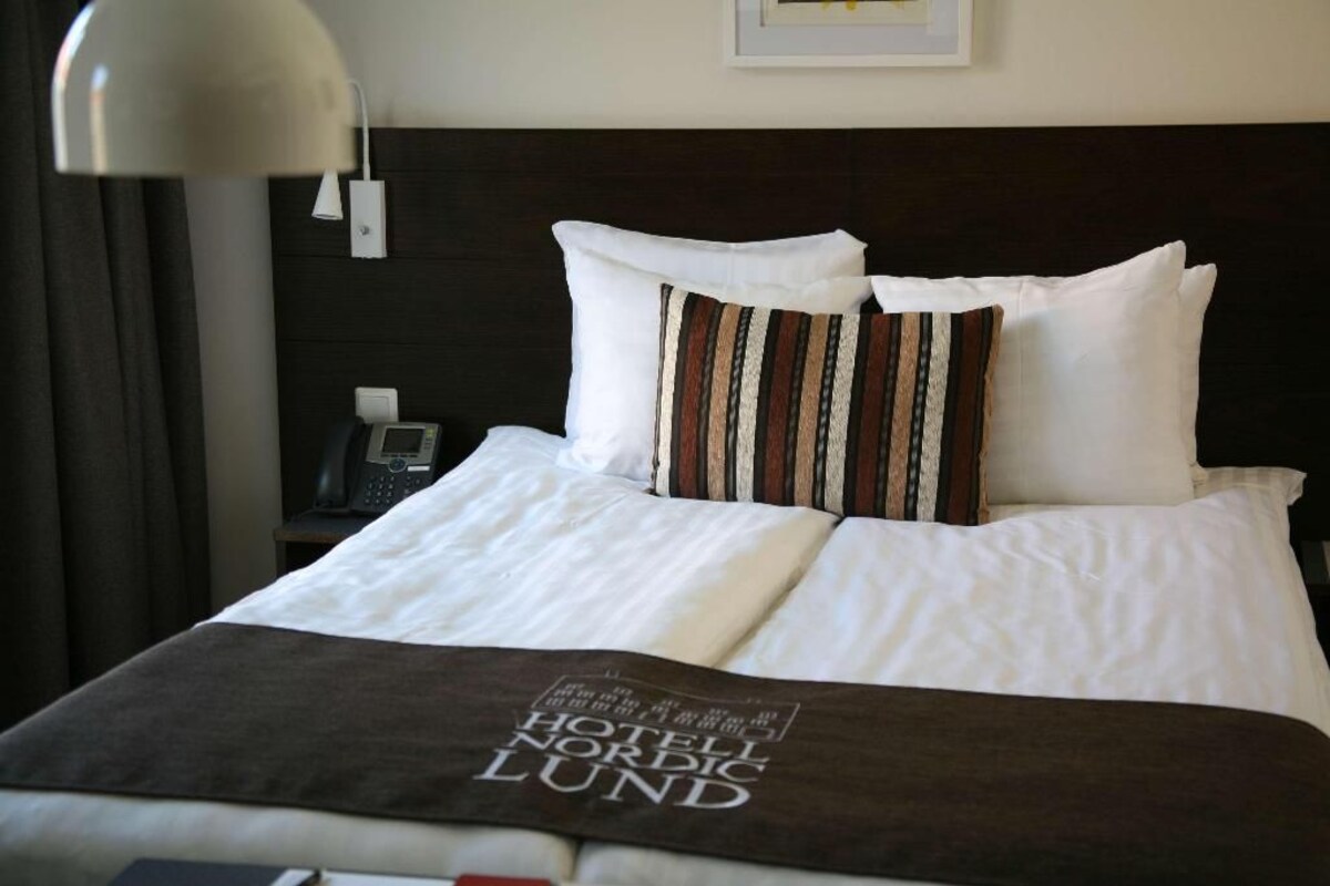Una cama bien tendida con una manta blanca y marrón, una almohada a rayas y una almohada con el logotipo del hotel.