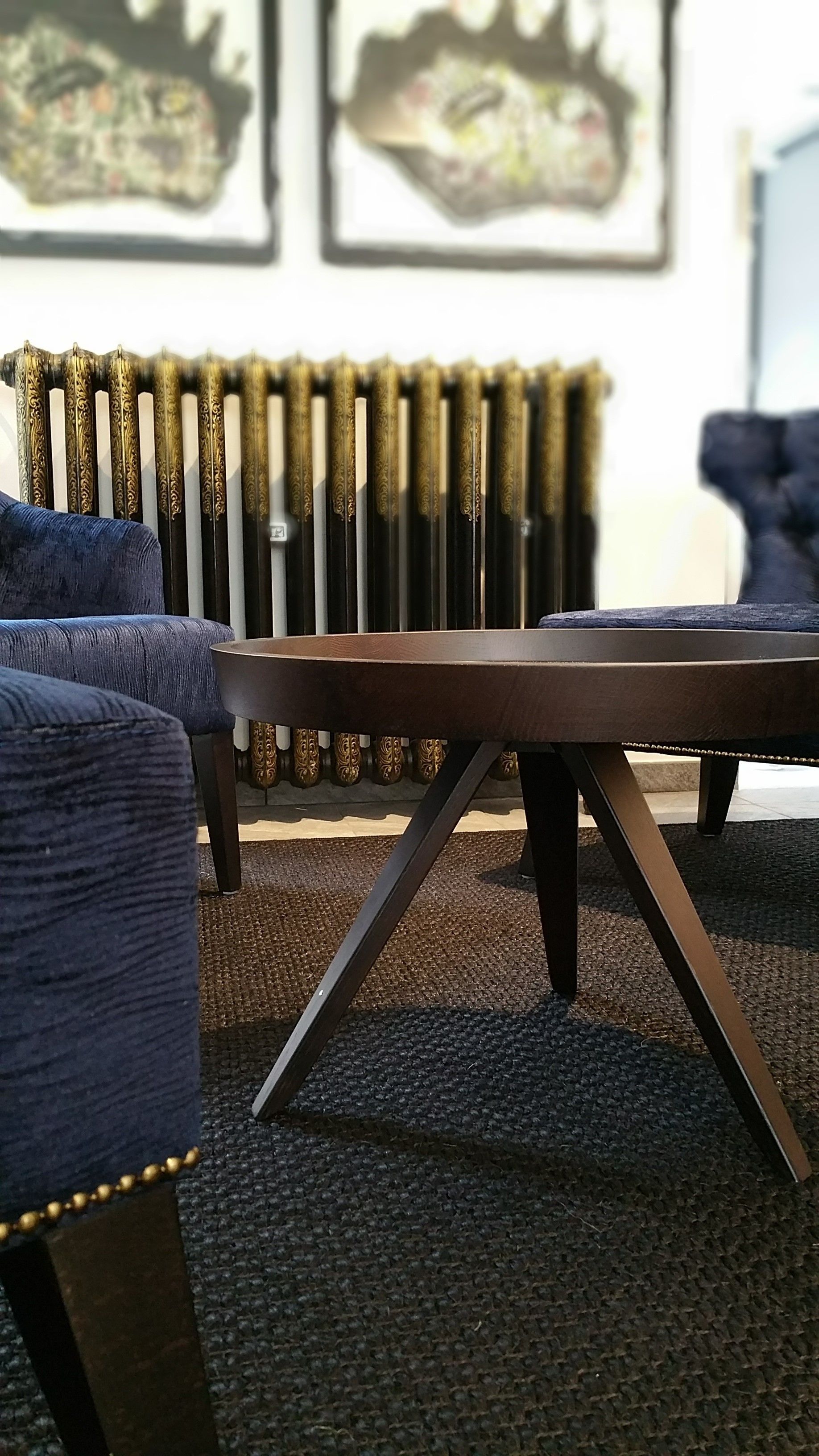 A modern living room with a dark brown coffee table, blue upholstered chairs, and a radiator with decorative patterns.