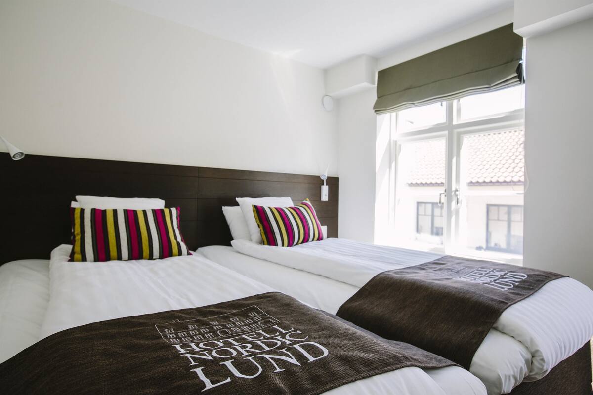 Quarto de hotel com duas camas, roupa de cama branca, almofadas listradas e janela com vista para edifícios.