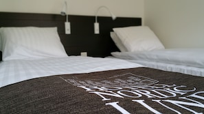 Quarto de hotel com duas camas, roupa de cama branca, cabeceira escura e manta com padrão de design nórdico.