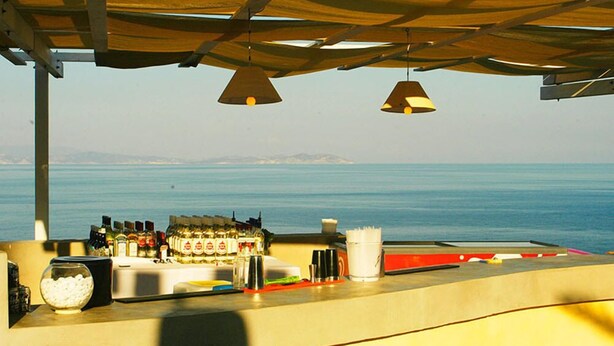 A bar area with a view of the sea, bottles of alcohol, and a white cup on the counter. - Cape Napos (Sifnos)