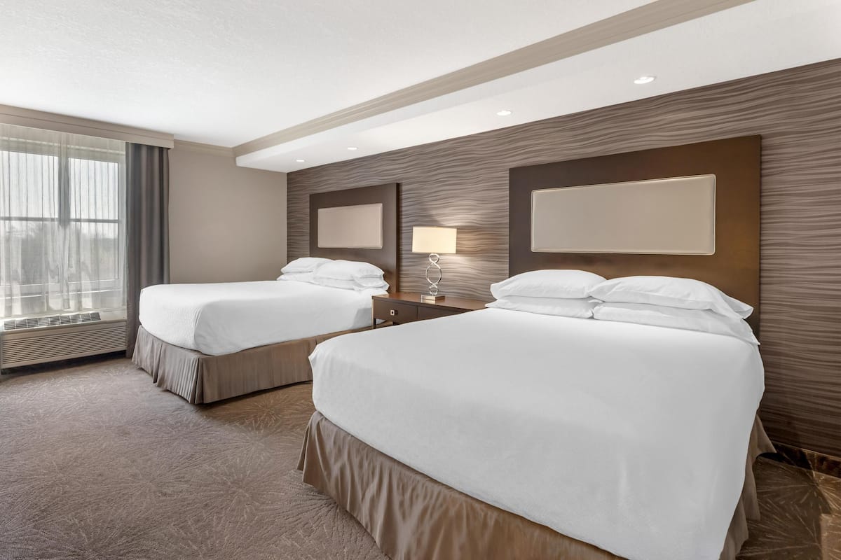 Habitación de hotel con dos camas, un ventanal con cortinas, una lámpara de pared y un panel decorativo de madera.