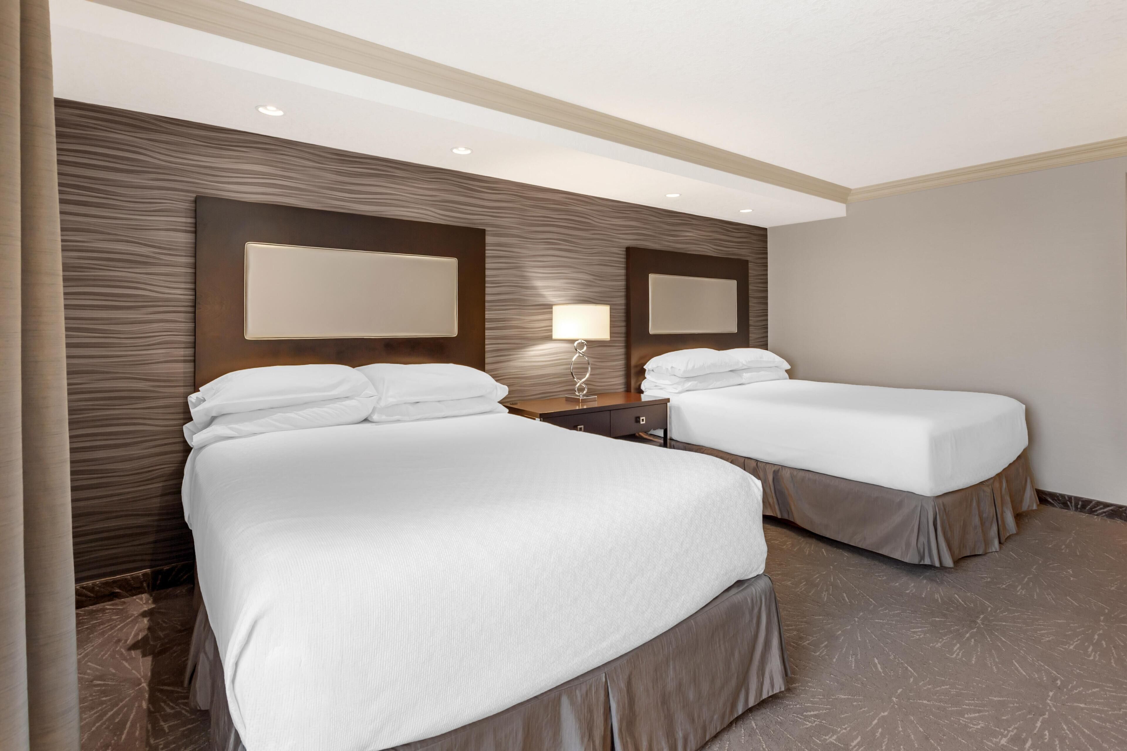 Quarto de hotel com duas camas, cabeceira de madeira, mesa de cabeceira, luminária e espelho.