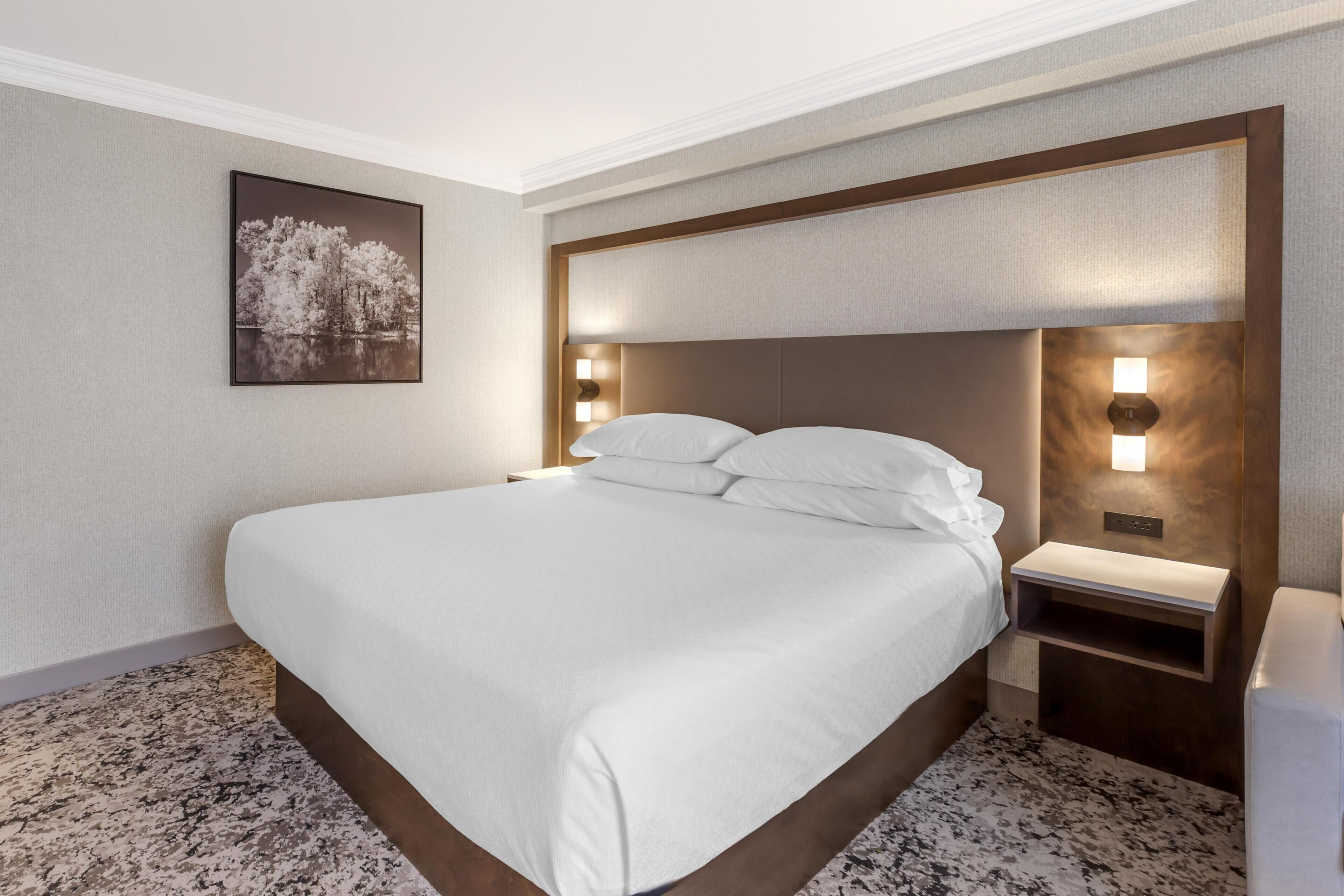 Una habitación de hotel con una cama grande, dos mesitas de noche, una lámpara de pared e imagen enmarcada en la pared.