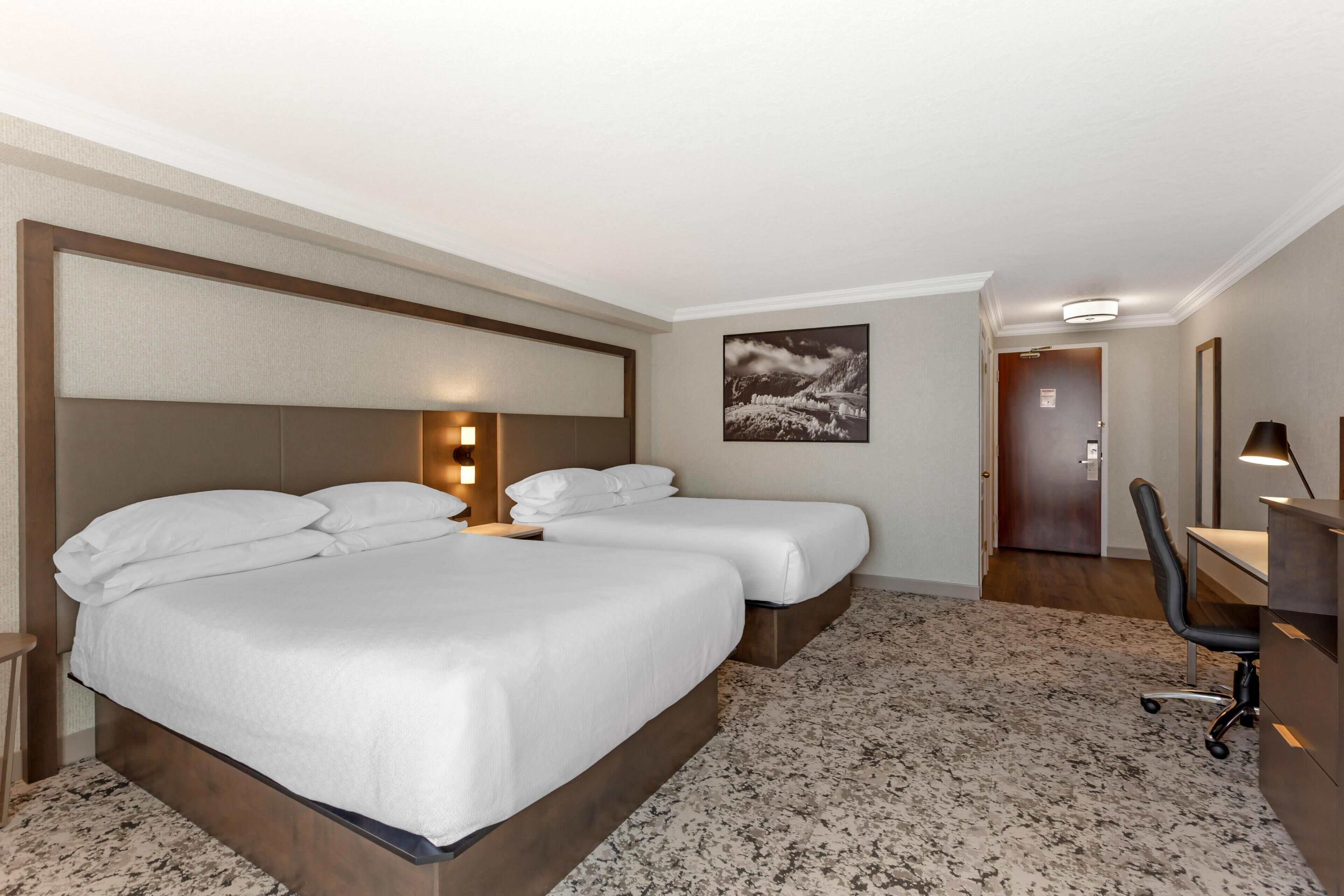 Une chambre d’hôtel avec deux lits, un bureau, une chaise, un tableau au mur et une porte donnant sur une autre pièce.