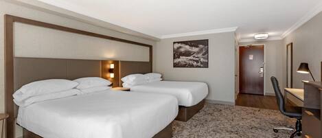 Une chambre d’hôtel avec deux lits, un bureau, une chaise, un tableau au mur et une porte donnant sur une autre pièce.