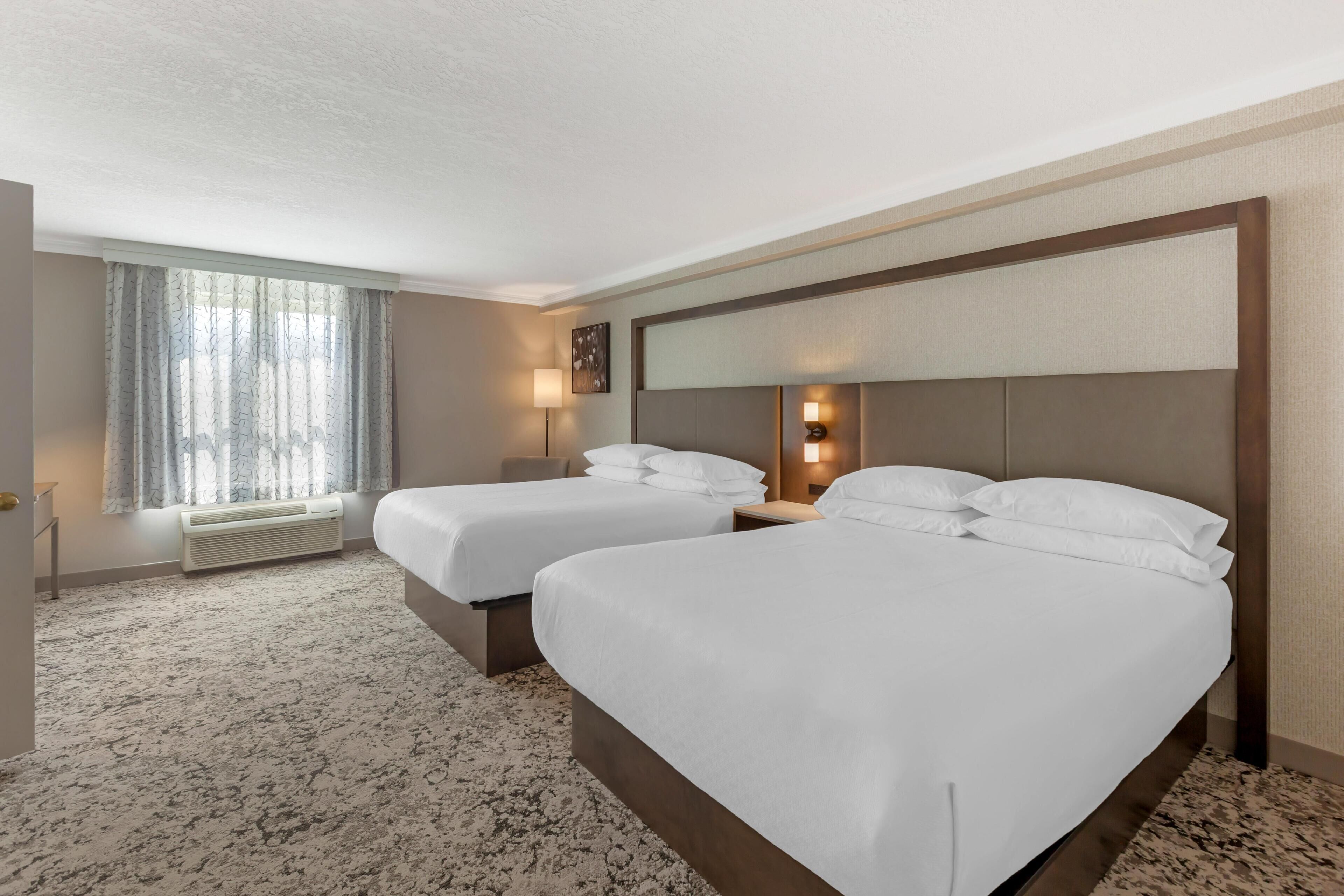 A hotel room with two beds, a large window with curtains, a wall-mounted lamp, and a carpeted floor.