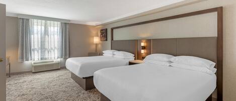 Habitación de hotel con dos camas, un ventanal con cortinas, una lámpara de pared y suelo alfombrado.
