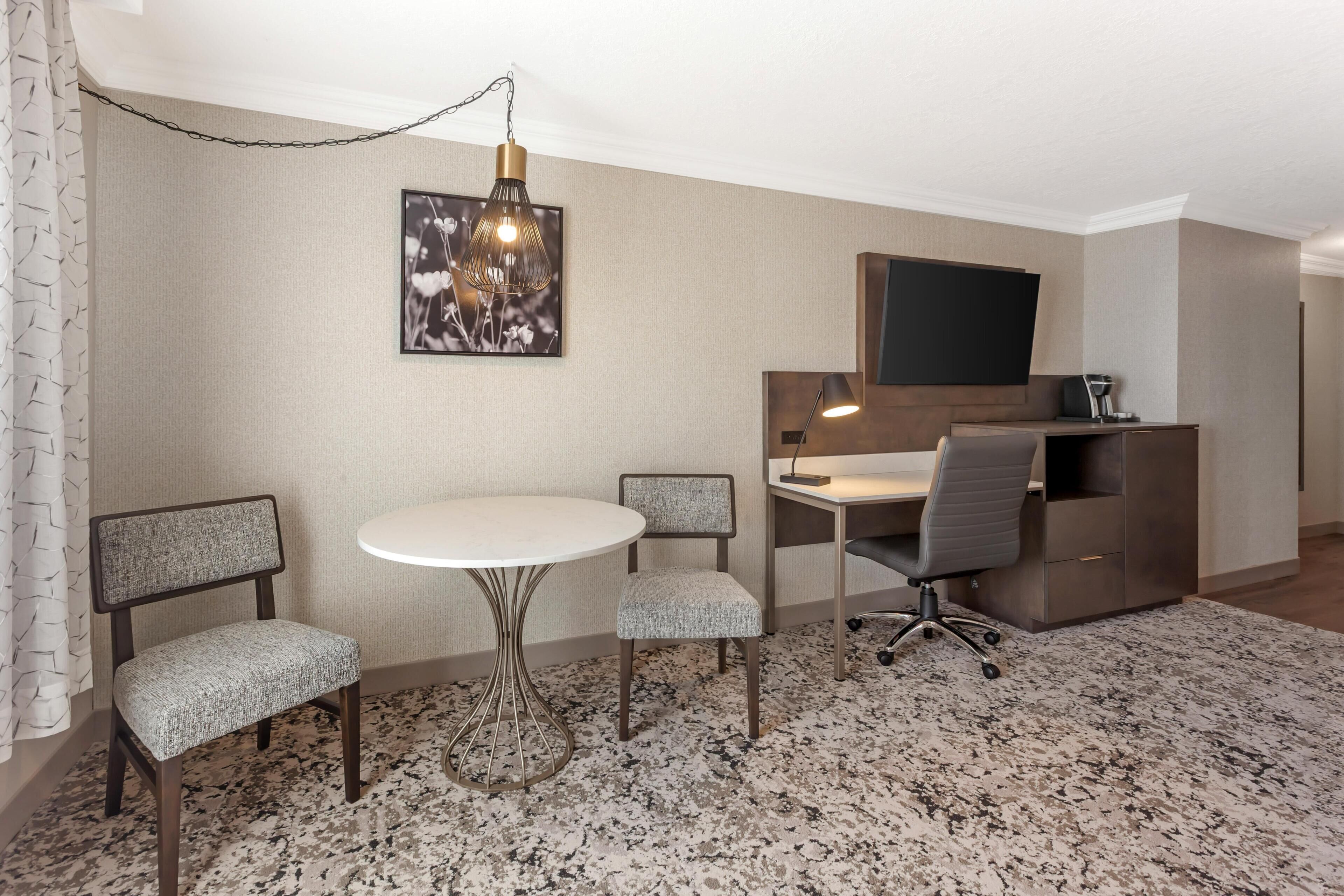 A hotel room with a round dining table, two upholstered chairs, a desk with a chair, a mounted flat-screen TV, and a patterned carpet.