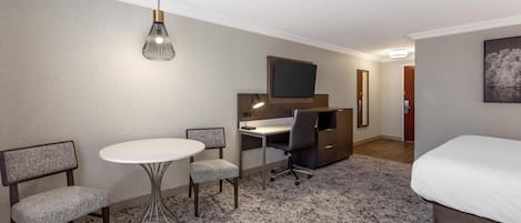 A hotel room with a bed, a desk with a chair, a small table, a TV, and a hanging light fixture.