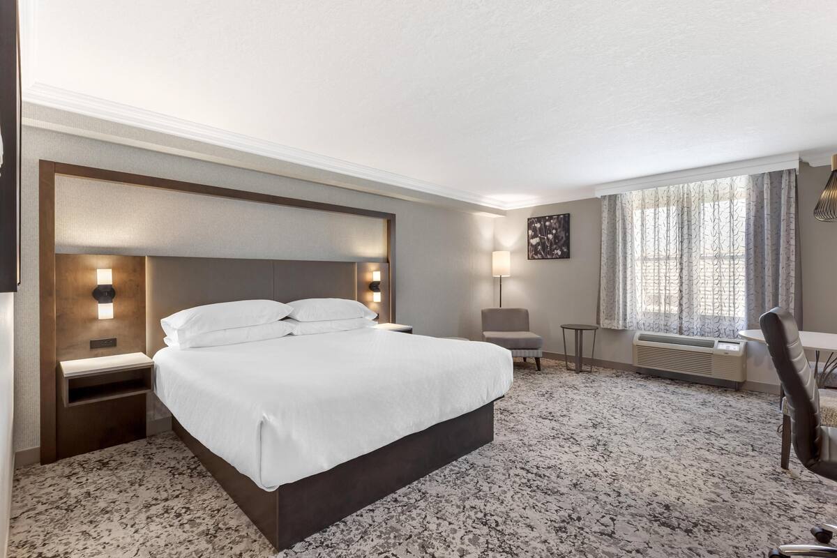 Habitación de hotel con una cama grande, mesitas de noche, una silla, una pequeña mesa y una ventana con cortinas.