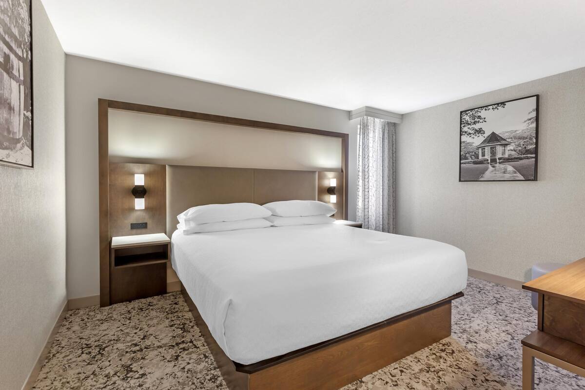 Una habitación de hotel con una cama grande, mesitas de noche, una mesita de madera, un cuadro en la pared y piso alfombrado.