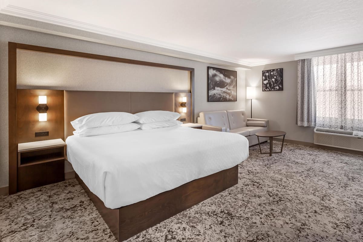 Una habitación de hotel con una cama grande, mesitas de noche, una silla, una pequeña mesa y una ventana con cortinas.