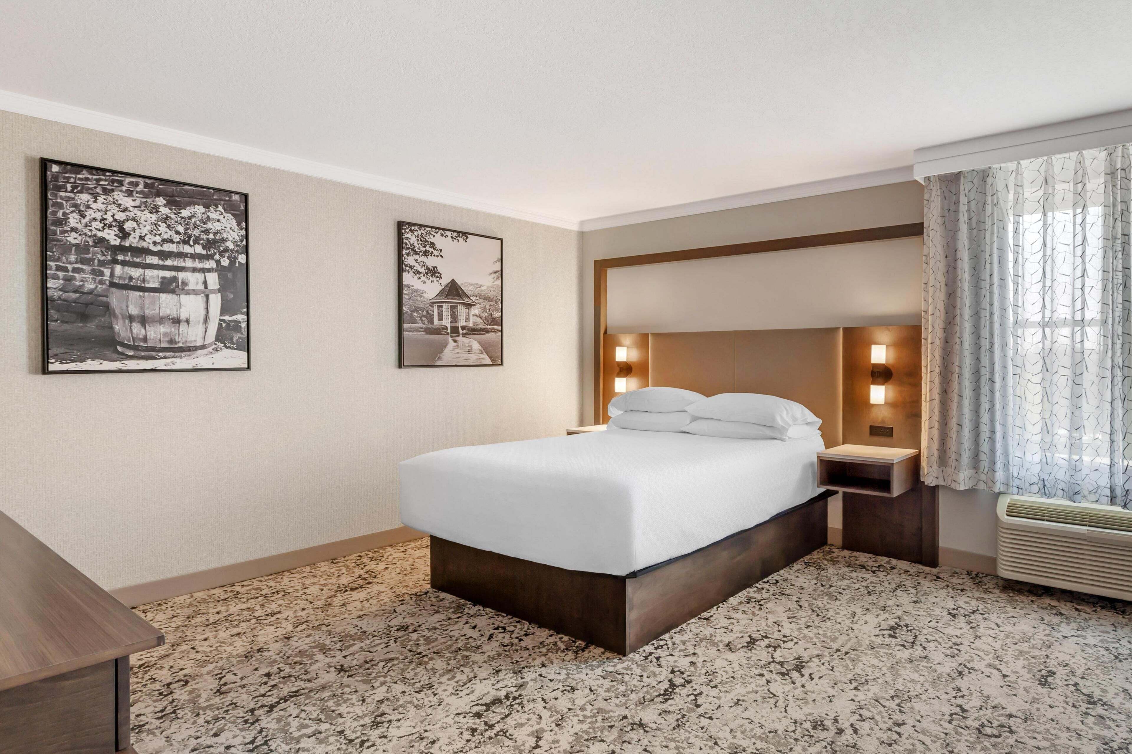 Una habitación de hotel con una cama, una mesita de noche, una lámpara de pared y dos cuadros enmarcados.