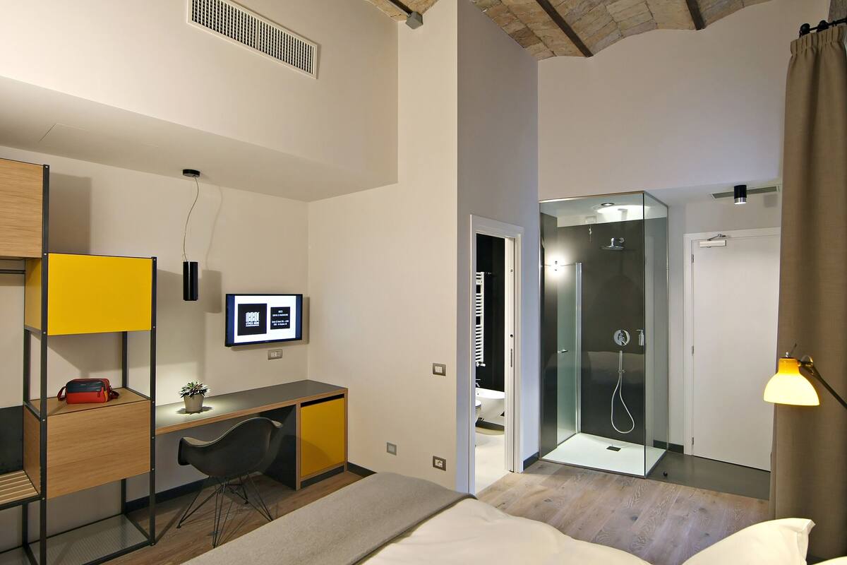 A modern hotel room with a wooden desk, a bed, a shower area, and a small seating area.