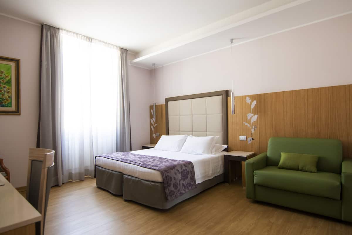 A hotel room with a bed, a green armchair, a desk, and a painting on the wall.