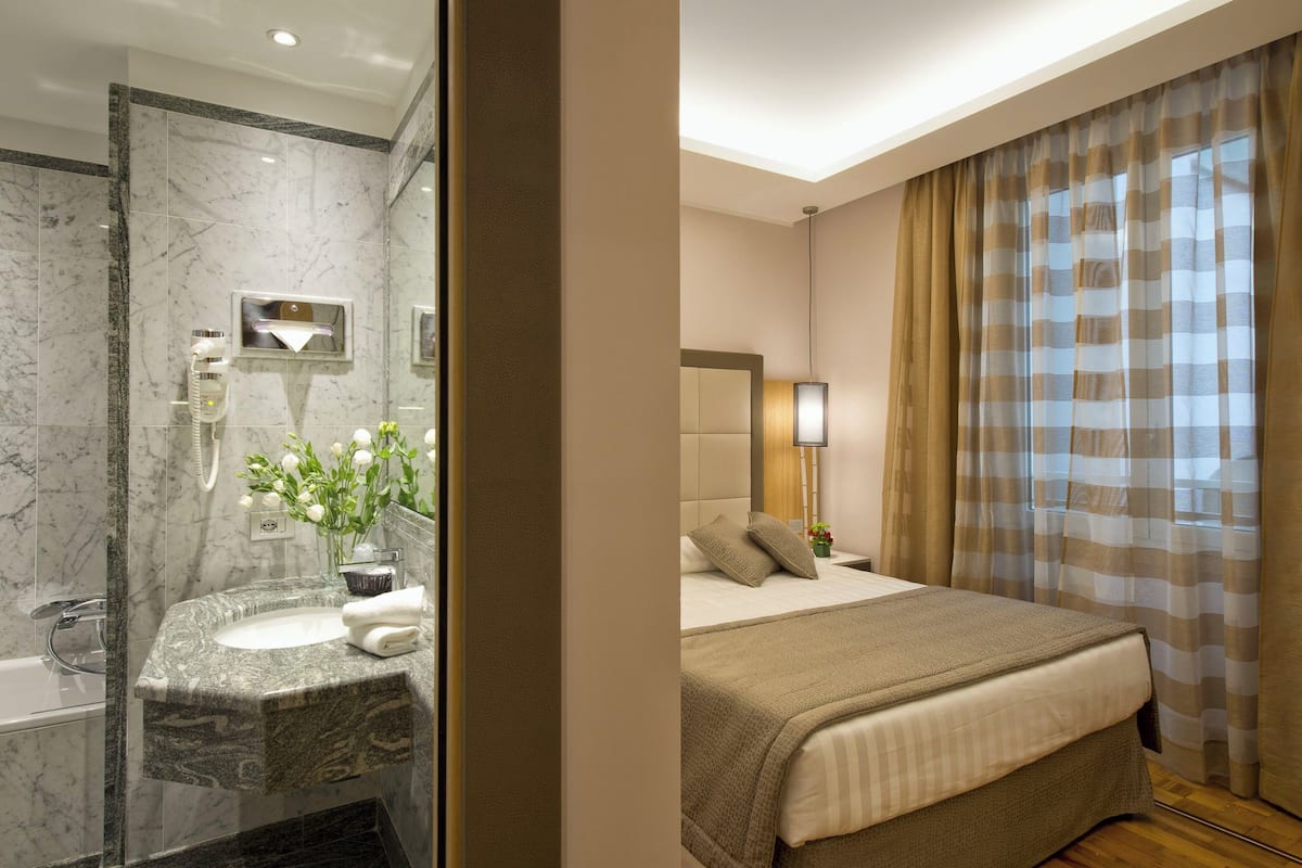 A hotel room with a marble bathroom, a bed with a beige bedspread, and a large window with curtains.