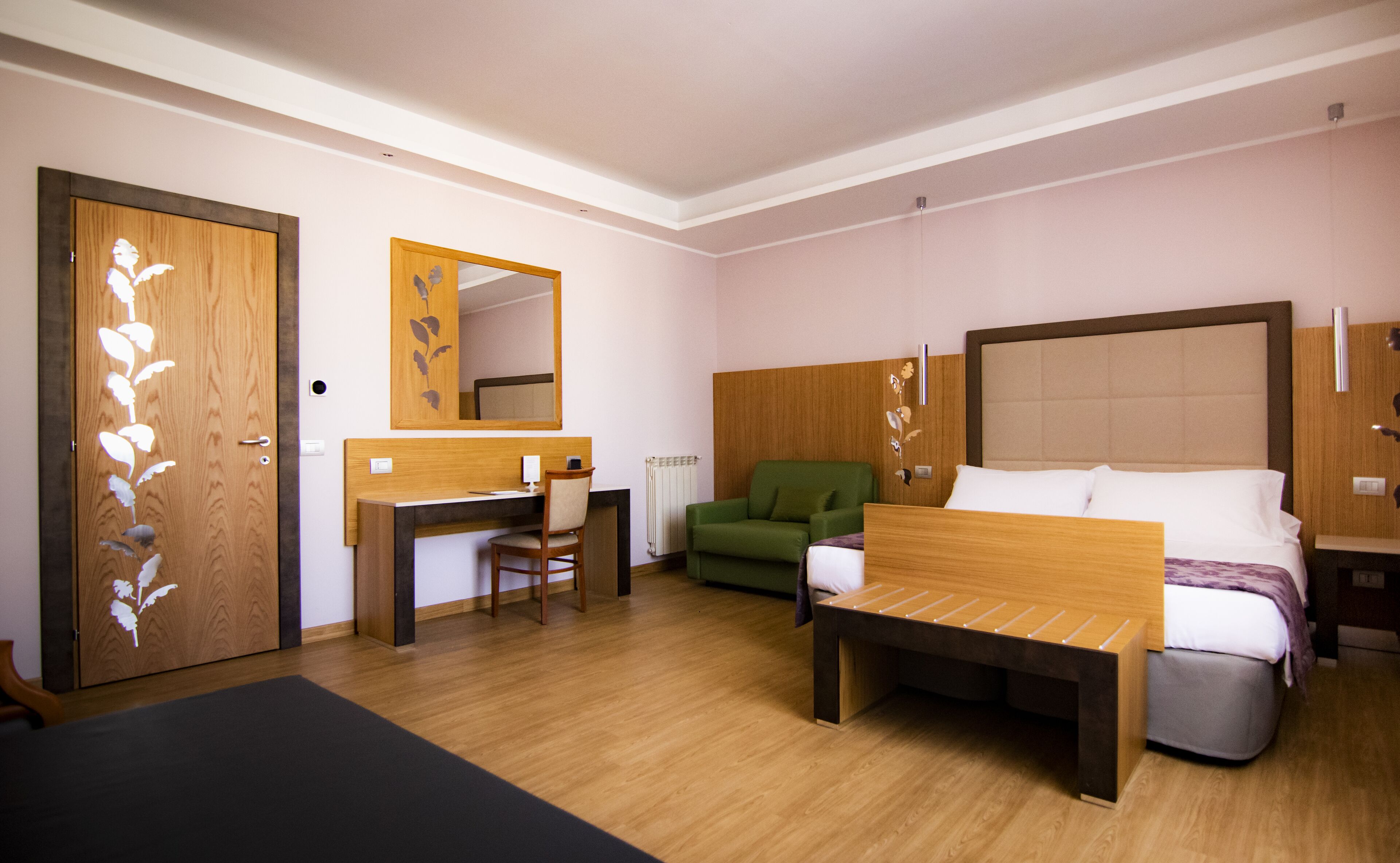 Habitación de hotel con un escritorio de madera, un sillón verde, una cama con cabecera de madera y un espejo grande en la pared.