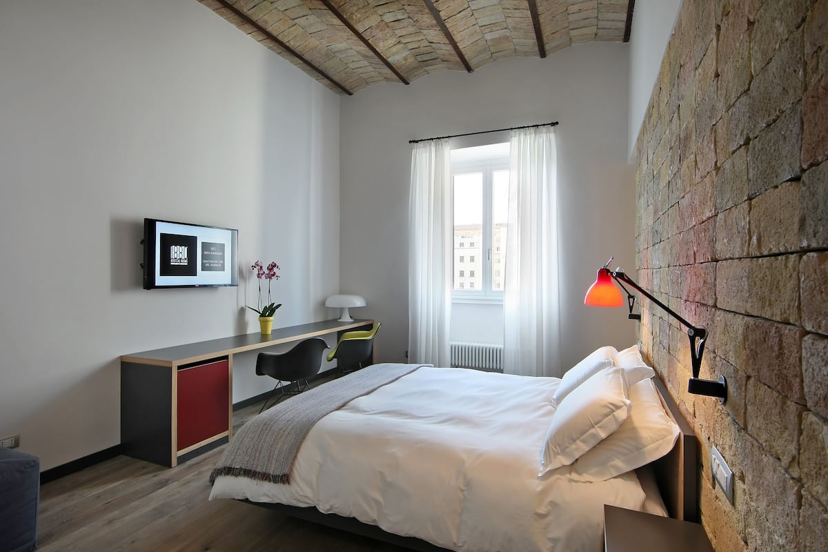 A modern bedroom with a bed, a desk, a chair, a lamp, and a window with curtains.